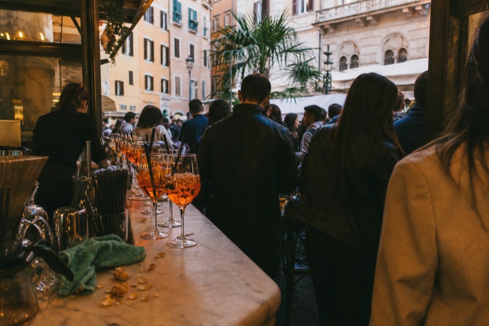A crowded bar counter in Rome at aperitivo hour with orange drinks lined up