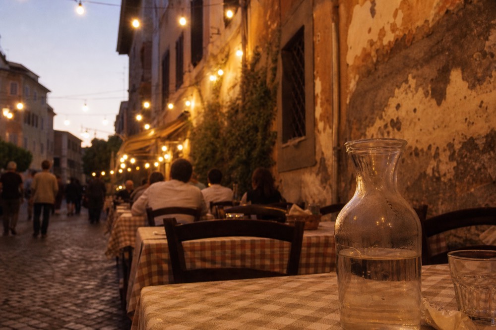 A busy Trastevere lane with restaurant tables lining the cobbles at dusk