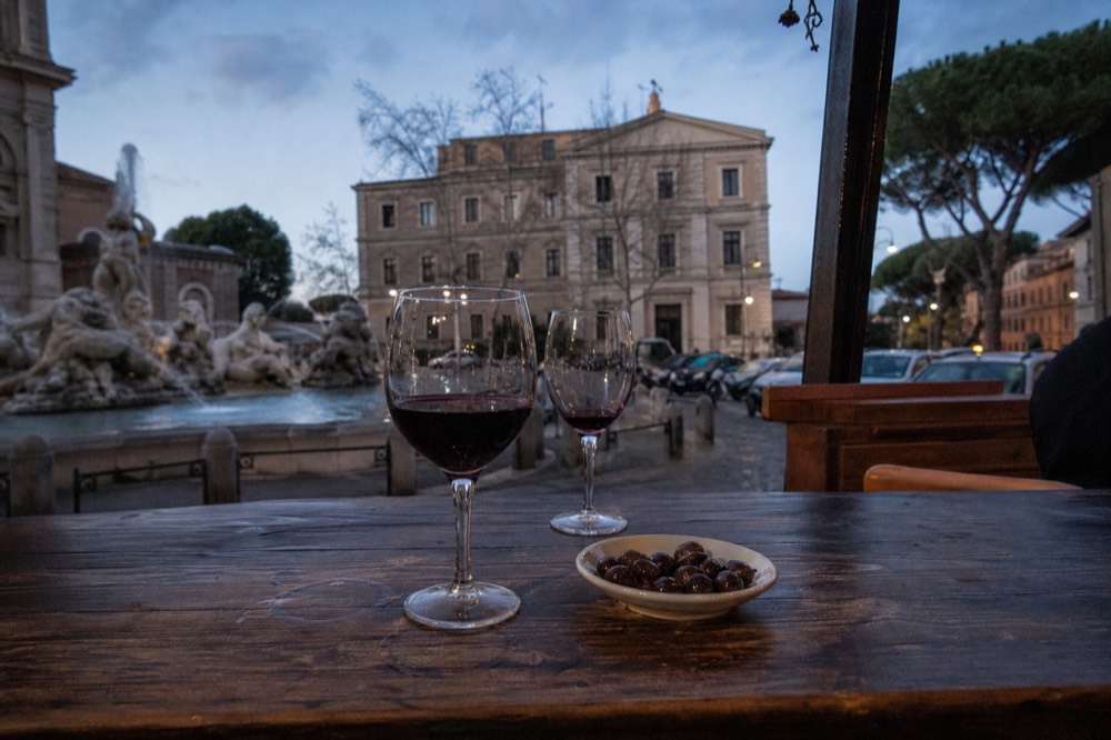 A glass of red wine and a small plate of olives on a wooden bar top