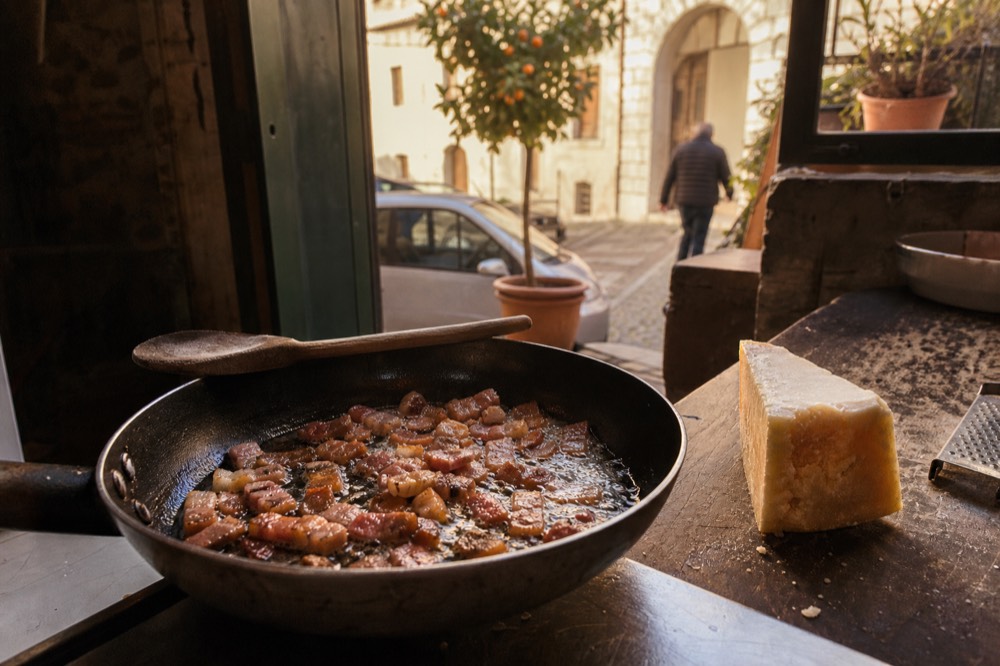 Guanciale cubes crisping in a pan beside a wedge of pecorino romano