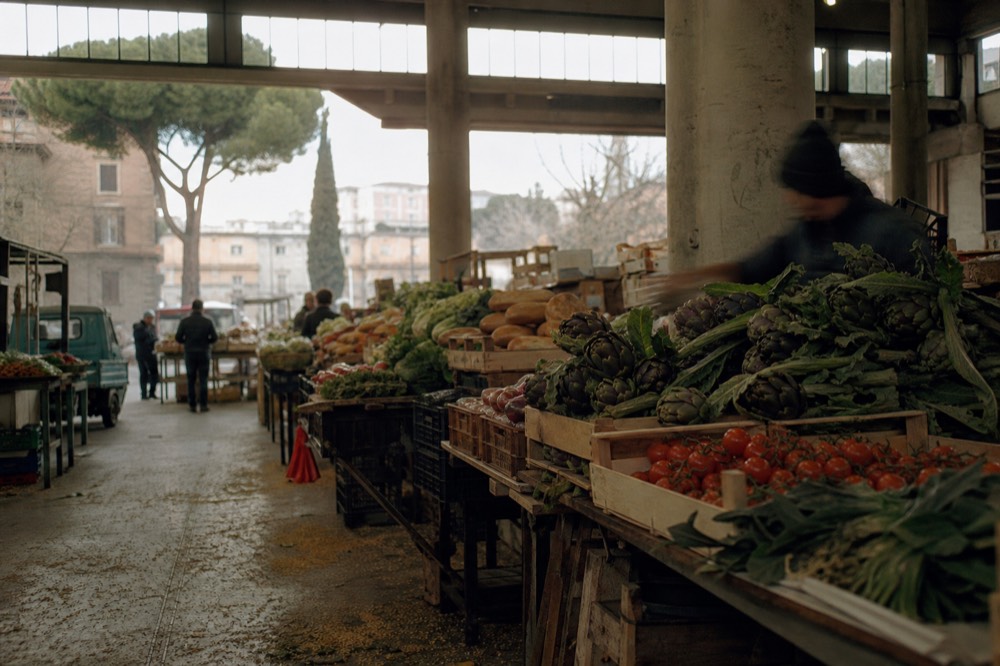Market stalls piled with Roman produce and bread inside a covered market hall