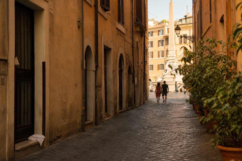 A quiet street leading away from a busy Roman piazza, with a restaurant sign in the distance